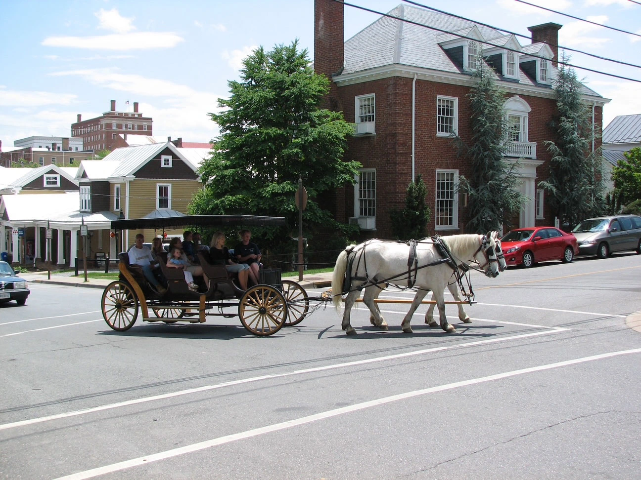 A horse-drawn carriage filled with passengers crosses a sunny street, surrounded by historic buildings and parked cars.